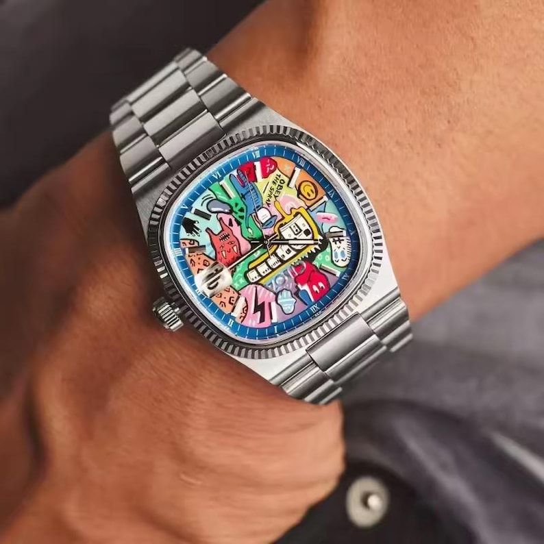 May include: A stainless steel wristwatch with a colorful, artistic dial. The watch face features a blue border and a vibrant design with various shapes and colors. The watch has a silver band and is worn on the wrist.