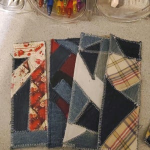 May include: A collection of handmade fabric bookmarks with a patchwork design. Each bookmark features a unique combination of denim, patterned fabric, and contrasting stitching. The color palette includes denim blue, red, white, and plaid patterns.