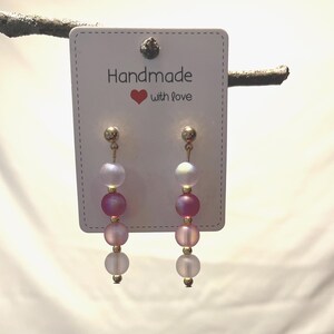 May include: Handmade earrings with a gradient of pink and white beads. The earrings are suspended from gold-colored posts and feature a heart and the words "Handmade with love" on the tag.