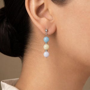 May include: A drop earring featuring a vertical arrangement of three smooth, round beads in light blue, pale yellow, and soft lavender, separated by small silver-toned metal spheres, suspended from a silver-toned post and butterfly back closure.