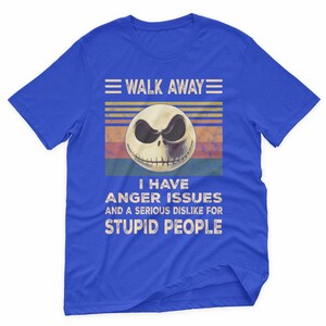 May include: Blue t-shirt with a graphic featuring a skull with a sinister expression. The text reads "WALK AWAY" above the skull and "I HAVE ANGER ISSUES AND A SERIOUS DISLIKE FOR STUPID PEOPLE" below.