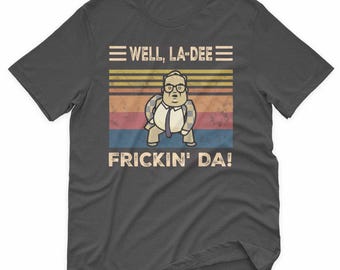 May include: Dark gray t-shirt featuring a cartoon illustration of a man in a suit with the text "WELL, LA-DEE" above and "FRICKIN' DA!" below. The background has horizontal stripes in shades of yellow, orange, red, and blue.