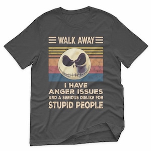 May include: Dark gray t-shirt featuring a graphic of a skull with the text "WALK AWAY" above and "I HAVE ANGER ISSUES AND A SERIOUS DISLIKE FOR STUPID PEOPLE" below. The skull has a sinister expression.