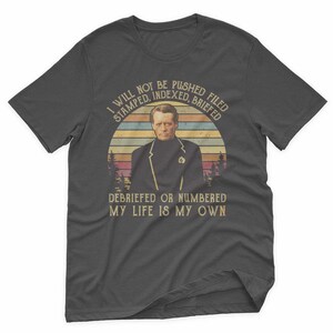 May include: Dark gray t-shirt featuring a vintage-style graphic. The graphic includes a portrait of a man in a black jacket against a sunset backdrop, with text that reads "I WILL NOT BE PUSHED, STAMPED, INDEXED, BRIEFED, DEBRIEFED OR NUMBERED, MY LIFE IS MY OWN."