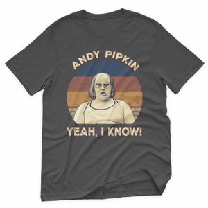 May include: Dark gray t-shirt featuring a retro-style graphic of a person with glasses and the text "ANDY PIPKIN" above and "YEAH, I KNOW!" below. The graphic is set against a circular background with blue, red, and orange stripes.