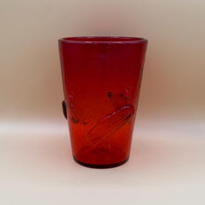 May include: A vibrant red glass tumbler featuring a distinct crackle texture throughout its surface. The vessel has a slightly tapered cylindrical shape with a smooth rim and a solid, weighted base, showcasing a deep crimson hue and translucent quality.