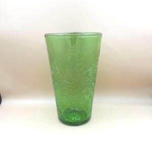 May include: A green glass tumbler with a crackle texture. The glass has a cylindrical shape and a thick base. The color is a vibrant green, and the texture gives it a unique, aged appearance. The glass is suitable for beverages.