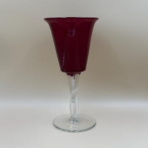 Könnte beinhalten: Ein Weinglas mit einer tief burgunderroten Kuppa und einem ausgestellten Rand. Der Stiel besteht aus klarem Glas, das zu einer Spirale gedreht ist und in einem flachen, runden Fuß aus klarem Glas endet, der dem Gefäß einen stabilen Stand verleiht.