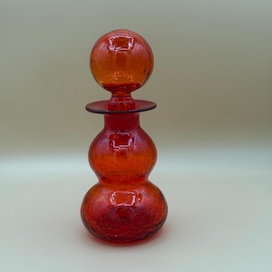 May include: A vibrant orange crackle glass decanter featuring a double-bulb body and a matching spherical stopper. The glass has a distinct webbed texture throughout, with a smooth, rounded neck and a wide, flat rim where the stopper rests.
