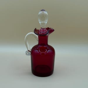 Peut inclure: Une cruche en verre rouge rubis avec une poignée et un bouchon en verre transparent. La cruche a un bord évasé et un corps bulbeux. La poignée est incurvée et fixée sur le côté. Le bouchon est allongé et s'insère dans le haut.