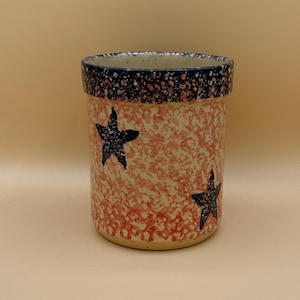 May include: A ceramic utensil holder with a speckled red and white pattern. The rim is dark blue with white speckles. Two dark blue stars are painted on the front. The holder is cylindrical.