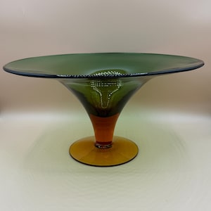 Könnte beinhalten: Eine dekorative Glasschale mit einer breiten, flachen grünen Oberseite und einem ausgestellten, kegelförmigen Sockel. Der Sockel geht von Grün zu Orange über und ruht auf einem kreisförmigen, bernsteinfarbenen Fuß. Die Schale wird wahrscheinlich zur Präsentation verwendet.