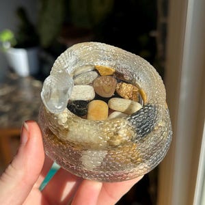 May include: A clear, coiled snake-shaped container filled with small, colorful stones. The container has a textured surface and a clear, protruding head. The stones are various colors, including beige, brown, and black.
