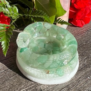Puede incluir: Un portavelas de cristal verde lleno de pequeñas piedras verdes. El portavelas está sobre una base blanca y tiene un lazo decorativo. Flores rojas y follaje verde en el fondo.