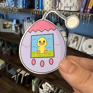 May include: A sticker in the shape of a retro digital pet device. The device is lavender with a pink top and three pink buttons. The screen shows a yellow duck on green grass. A white chain and circle are attached.