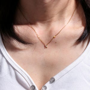 以下が含まれることがあります： 小さな輝く文字「X」のペンダントと小さなハートチャームが付いた繊細なゴールドネックレス。白いトップスに合わせて着用されており、シンプルでエレガントな印象です。チェーンは細く、ペンダントは小さいです。