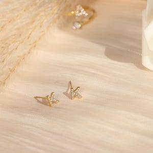 Peut inclure: Boucles d'oreilles dorées en forme de la lettre « K », incrustées de petits cristaux transparents. Les boucles d'oreilles sont présentées sur une surface beige clair, avec une bague assortie et une fleur blanche en arrière-plan.