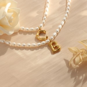 May include: Two pearl necklaces with gold letter charms, one with a "C" and the other with a "B". A cream-coloured rose and dried foliage are in the background. The necklaces are on a light peach-coloured surface.