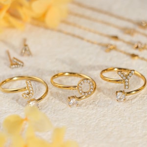 Puede incluir: Anillos dorados con las letras "J", "O" e "Y" incrustadas con pequeños cristales transparentes. Se ven otras joyas, incluidos pendientes y collares, en el fondo.