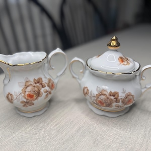 May include: A white porcelain creamer and sugar bowl set. The set features a floral design in shades of brown and orange, with gold trim. The sugar bowl has a lid with a gold finial.