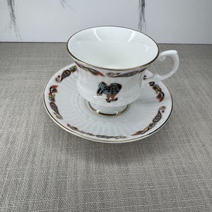 May include: White teacup and saucer set with gold trim and a decorative pattern of colorful paisley and a figure. The teacup has a handle and sits on a matching saucer. The set is likely porcelain.