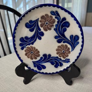 May include: A round ceramic plate with a cream-colored base, featuring a hand-painted design of three brown flowers and dark blue swirling leaf patterns. The plate has a scalloped edge with a thick dark blue border and rests on a black stand.