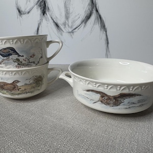 May include: A set of cream-colored ceramic serving dishes. Two handled soup bowls are stacked, each with a different bird illustration. A larger oval serving dish with handles features a bird in flight. All pieces have a decorative rim.
