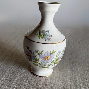 May include: A small white porcelain vase with a narrow neck and a flared rim. The body features a delicate floral pattern with light purple and blue flowers, green leaves, and thin gold trim accents around the rim, base, and middle section.