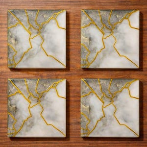 May include: Four square coasters with a white and gray marble design, accented with gold-colored lines. The coasters are arranged on a wooden surface, showcasing a modern and elegant aesthetic.