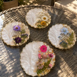 Puede incluir: Cuatro posavasos redondos de resina con bordes festoneados y una base blanca nacarada. Cada posavasos está decorado con flores prensadas, follaje verde y copos de pan de oro dispuestos en un patrón semicircular en un lado de la superficie.