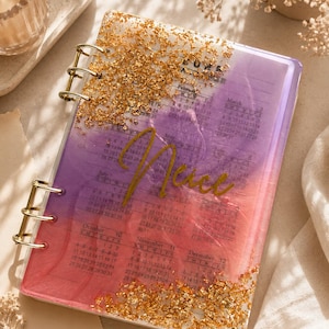 May include: A spiral-bound planner with a translucent cover featuring a purple and pink gradient background, gold foil flakes at the top and bottom, and the word "Nice" in gold script. The cover displays a full-year calendar grid.