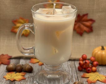 May include: A candle in a clear glass mug with a handle, filled with a creamy wax and a wick. The candle is surrounded by fall-themed decorations, including artificial leaves, acorns, and a small pumpkin.