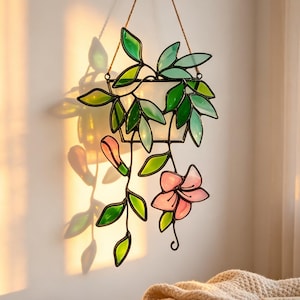 Peut inclure: Un attrape-soleil en vitrail représentant une jardinière suspendue avec des vignes traînantes, des feuilles vertes et une fleur rose. L'objet est assemblé avec des soudures en métal sombre et suspendu par un cordon fin pour refléter la lumière.