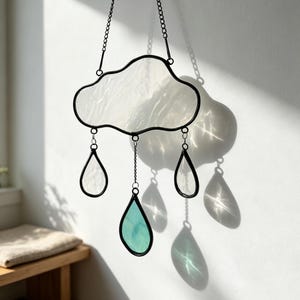 Peut inclure: Un attrape-soleil en vitrail en forme de nuage avec trois pendentifs en forme de goutte. Le nuage et les deux gouttes extérieures sont en verre blanc texturé, tandis que la goutte centrale est en verre bleu sarcelle translucide, le tout cerclé de noir.