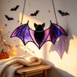 May include: A stained glass bat suncatcher featuring a black body and iridescent purple and blue wings. The bat is suspended by two black metal chains attached to the top of its wings, creating a decorative hanging piece with dark outlines.