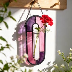 May include: A wall-hanging stained glass propagation station featuring an oval black metal frame with purple and pink glass panels. A clear glass test tube is suspended in the centre, holding a single green stem with two vibrant red flowers.