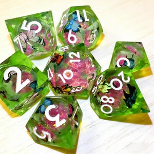 May include: A set of seven polyhedral dice for tabletop games. Each die is transparent green with white numbers and contains butterflies and pink glitter. The dice include a D4, D6, D8, D10, D12, D20, and a percentile die.