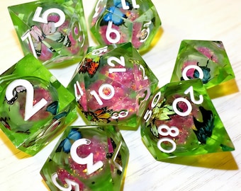 Op de afbeelding: Een set van zeven polyhedrale dobbelstenen voor tabletopspellen. Elke dobbelsteen is transparant groen met witte cijfers en bevat vlinders en roze glitter. De dobbelstenen bevatten een D4, D6, D8, D10, D12, D20 en een percentieldobbelsteen.