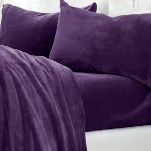 May include: A close-up of a bed with a deep purple fleece blanket, pillowcases, and fitted sheet. The bedding is soft and plush, with a velvety texture. The pillows are rectangular and the blanket is draped over the bed.