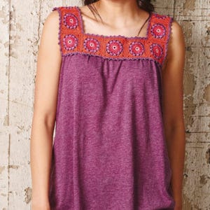 Puede incluir: Una blusa sin mangas morada con cuello cuadrado. El cuello presenta un borde de ganchillo en naranja y rosa, con un diseño floral. La blusa está hecha de una tela suave y texturizada.
