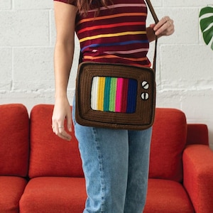 May include: A brown crochet crossbody bag shaped like a vintage television. The screen displays a colorful array of vertical stripes. The bag has two white circular buttons on the side and a brown strap.