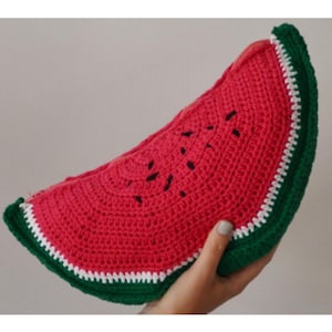 Puede incluir: Una rebanada de sandía hecha a mano con ganchillo en tonos rojos, verdes y blancos. La sandía tiene detalles de semillas negras y una corteza verde con bordes blancos. El artículo se sostiene en una mano.
