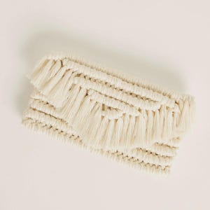 May include: Cream-coloured macrame clutch bag with a rectangular shape. The bag features a woven design with layered fringe detailing across the top. The neutral colour and textured surface create a bohemian aesthetic.
