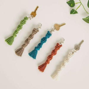 May include: Five macrame keychains in shades of green, brown, blue, orange, and white. Each keychain has a knotted design with a tassel at the bottom and a silver-colored clasp. Keys are attached to each keychain.