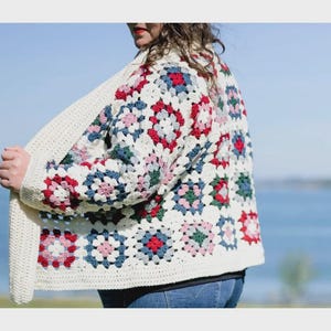 May include: A cream-colored, open-front crochet cardigan with a colorful floral pattern. The pattern features red, blue, pink, and green squares on a white background. The cardigan is worn over a dark top and blue jeans.