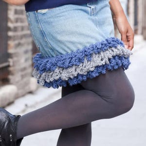 May include: A denim skirt with a blue and gray crocheted ruffle trim. The skirt is worn with gray tights and black boots. The crocheted trim adds a unique, handmade touch to the skirt.