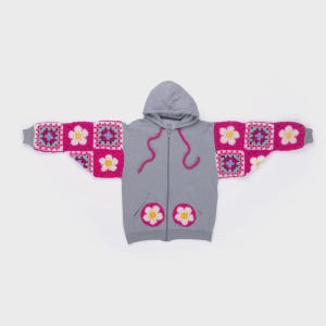 May include: A gray zip-up hoodie with a hood and two front pockets. The sleeves are adorned with colorful crochet squares in pink, purple, and white floral patterns. The hood drawstring is pink, and the pockets feature white and yellow flower appliqués.
