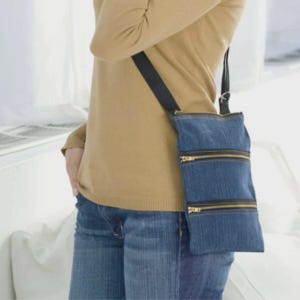 Op de afbeelding: Een denim crossbody tas met drie compartimenten met ritssluiting en een zwarte band. De tas is gemaakt van blauwe denim stof en heeft goudkleurige ritsen. De tas wordt over de schouder gedragen.