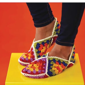 May include: A pair of colorful, handmade crochet slippers. The slippers feature a patchwork design with squares of red, orange, yellow, green, blue, and purple. The edges are trimmed with white yarn. The slippers are on a yellow surface.