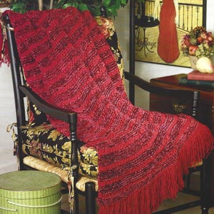 Pode incluir: Uma manta vermelha de malha com franjas está sobre uma cadeira de balanço de madeira preta com uma almofada com estampa floral. A manta tem um design listrado texturizado. Uma caixa de chapéu listrada em verde e branco está em primeiro plano.